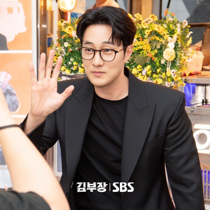 So Ji-sub in a black suit and glasses waves at the camera, displaying a friendly expression. He is surrounded by colorful flowers and promotional materials in the background.