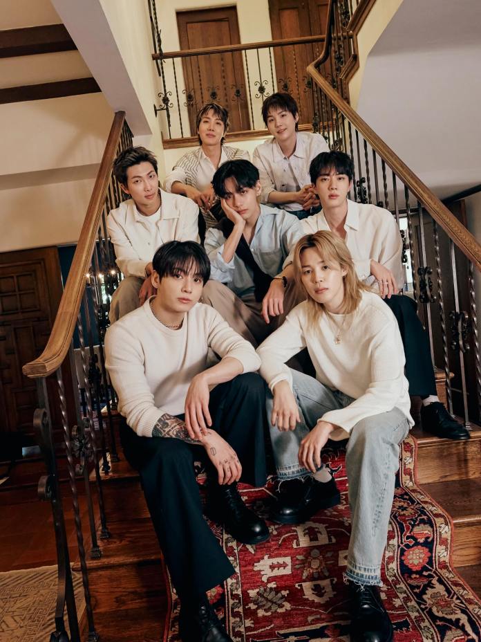 BTS members sitting together on a staircase, dressed in casual and stylish outfits, with a decorative rug underneath and wooden doors visible in the background.