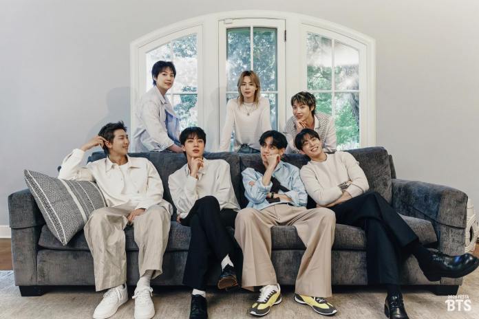 BTS members sitting on a gray couch in a bright room with a large window, wearing casual and stylish clothing.