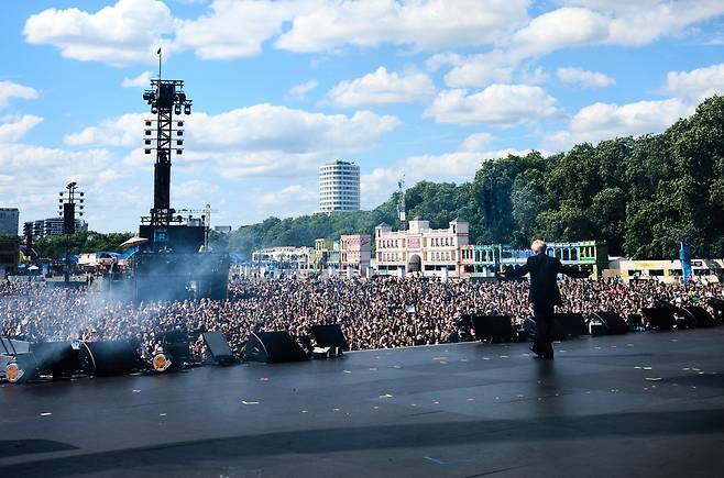 Taemin on stage facing a large crowd at a music festival, with a bright blue sky and fluffy clouds in the background.