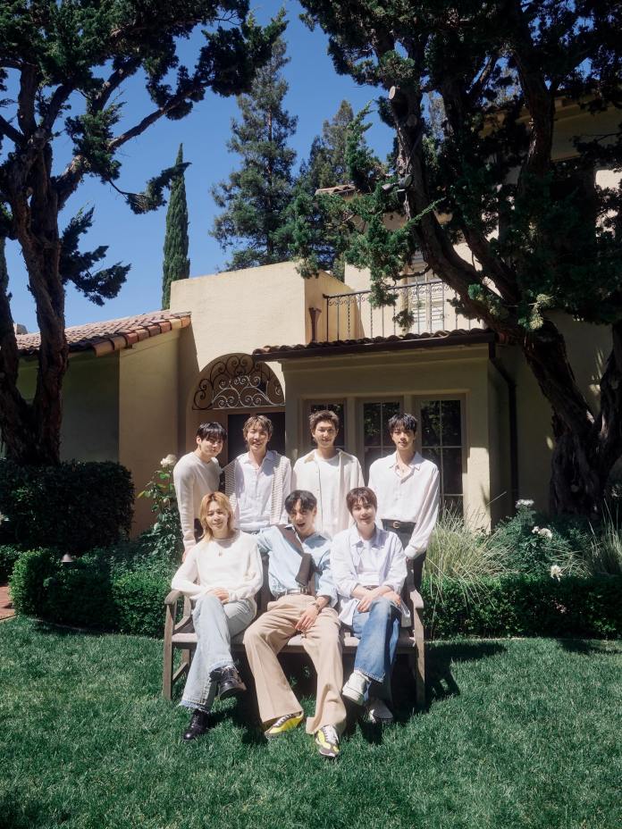 BTS members posing outdoors on a sunny day in a garden, with lush greenery and a house in the background.