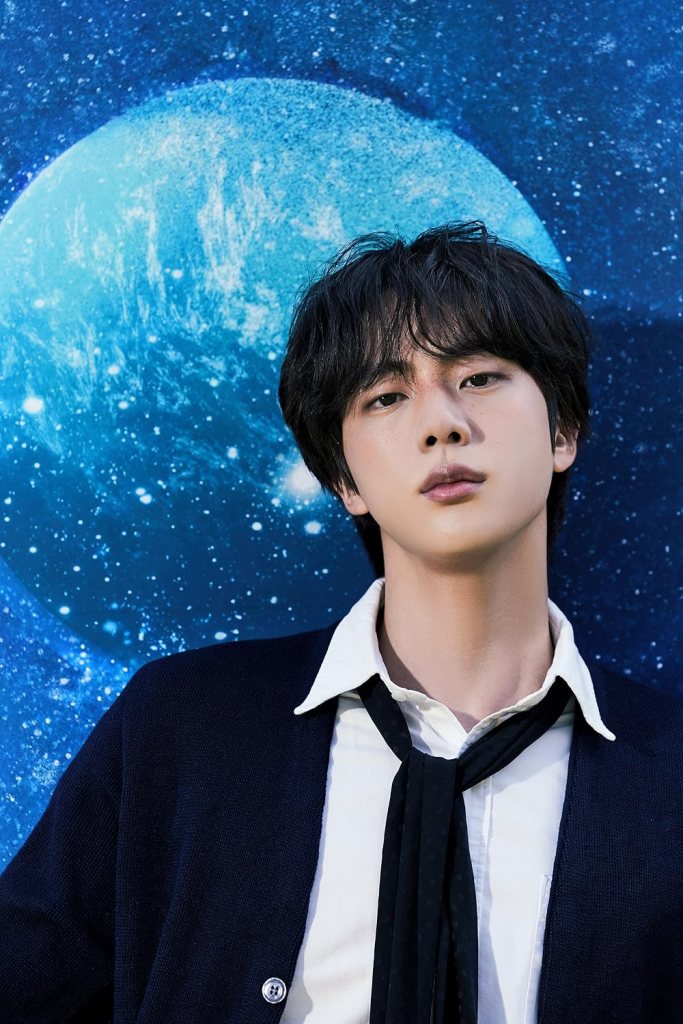 Jin with dark hair wearing a white shirt and dark cardigan stands in front of a blue, starry background with a planet.