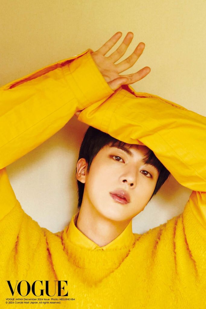 Jin wearing a bright yellow sweater, gazing at the camera with a soft expression. Their hand is raised near their face, set against a light background with the text 'VOGUE' in the corner.