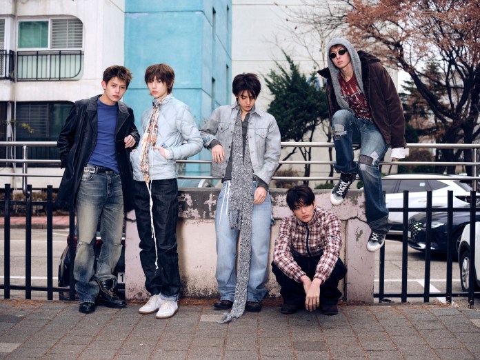CORTIS members posing together in urban attire, standing and sitting at a railing in a city environment with buildings and parked cars in the background.