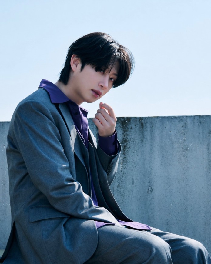 Heechul with long black hair, wearing a gray suit and a purple shirt, seated thoughtfully against a concrete wall under a clear blue sky.