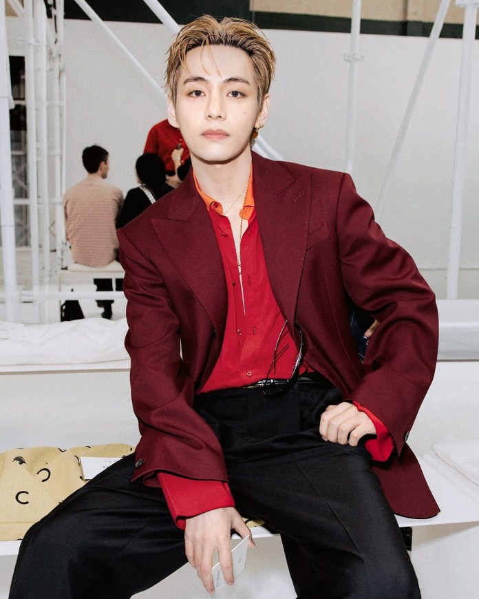 V sitting casually, wearing a burgundy suit jacket over a red shirt, with black pants, and holding a smartphone, in a modern indoor setting.