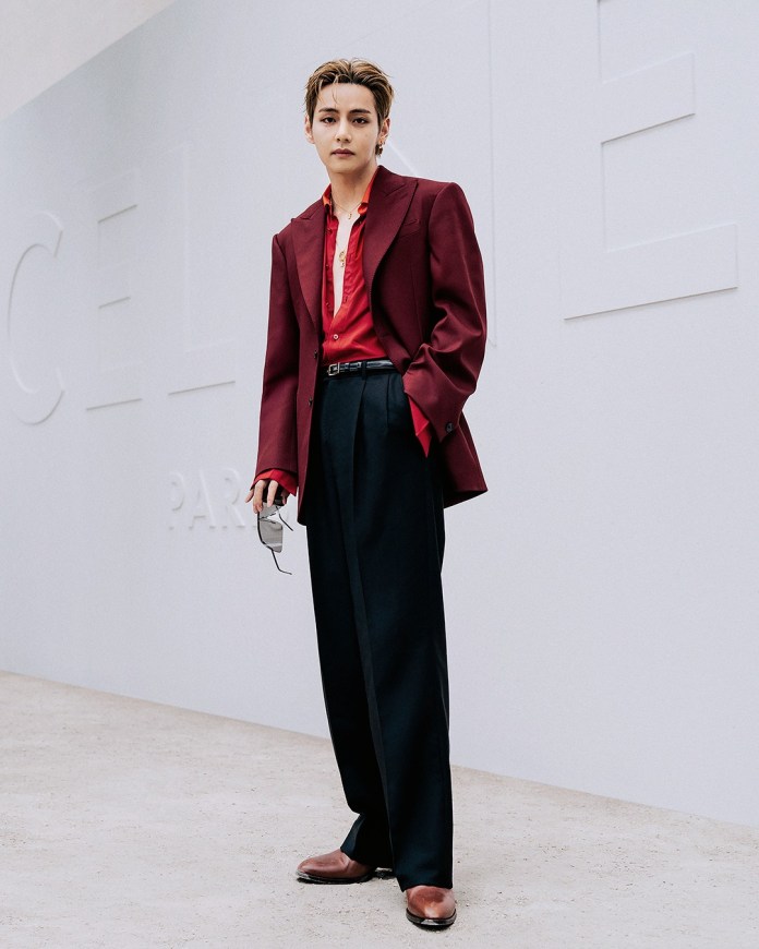 V stands confidently in a stylish outfit featuring a maroon blazer and a red shirt, paired with sleek black trousers and brown shoes. The background showcases a minimalist design with embossed letters.