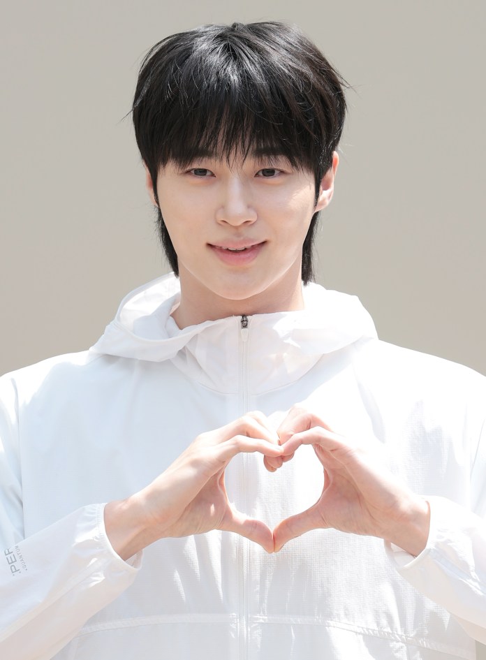 Byeon Woo-seok in a white jacket making a heart shape with their hands, smiling at the camera.