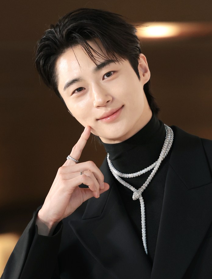 Byeon Woo-seok wearing a black turtleneck and a stylish jacket, adorned with a sparkling necklace, smiles and poses thoughtfully with one hand on his chin.