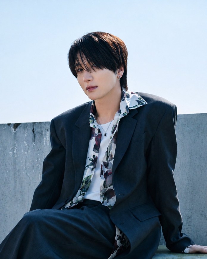 Leeteuk with dark hair sitting on a ledge against a blue sky, wearing a dark suit and a floral shirt.