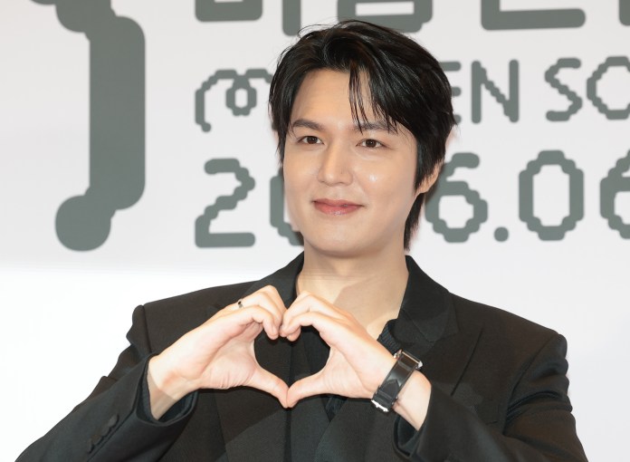 Lee Min-ho in a black suit making a heart shape with his hands, smiling at an event.