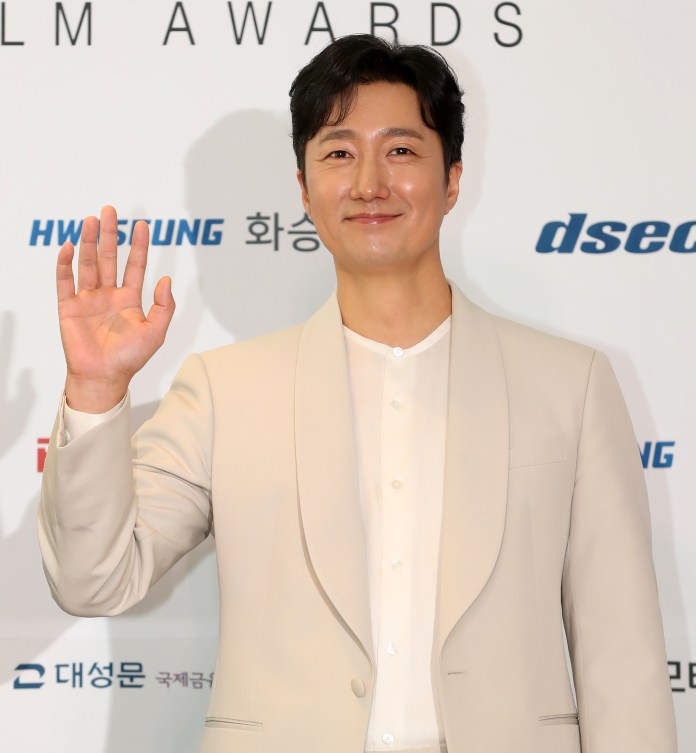 Park Hae-il wearing a beige suit and white shirt is smiling and waving at the camera at a film awards event.
