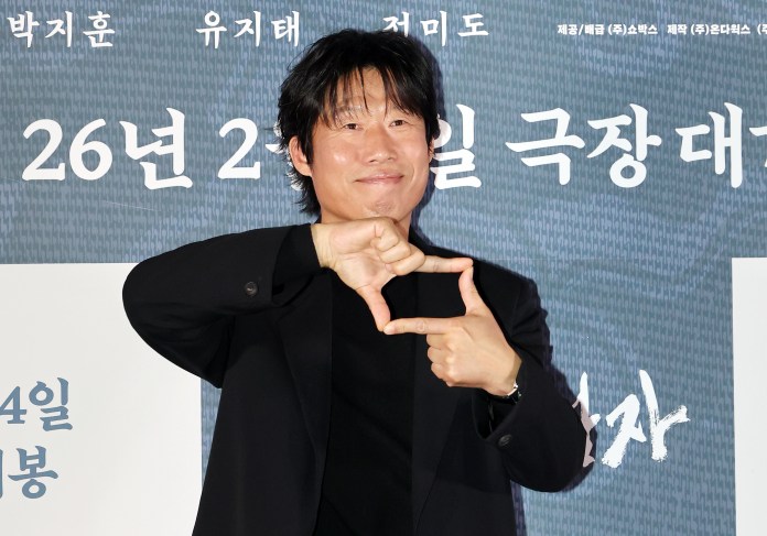 Yoo Hae-jin in a black jacket is making a hand gesture resembling a camera frame, standing in front of a backdrop with Korean text.