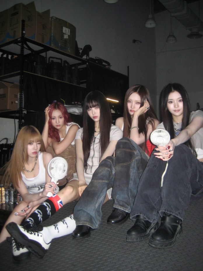 Keyveatz sitting together on a textured surface, dressed in casual outfits, with varying hairstyles. Some are holding small fans, and the background features shelves with boxes.