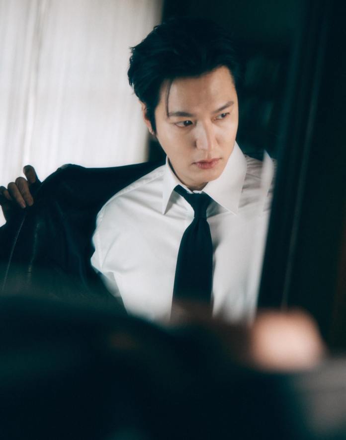 Lee Min-ho in a white shirt and black tie adjusts his jacket while looking into a mirror.