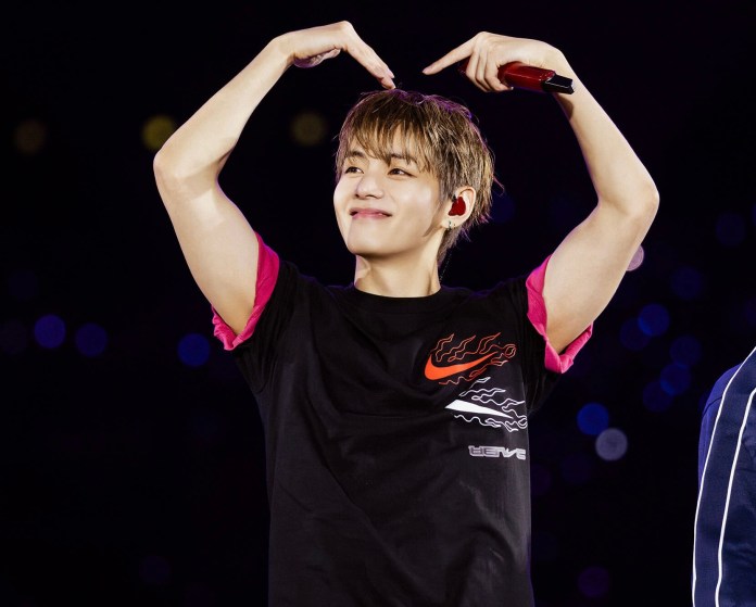 V smiling and making a heart shape with his hands, wearing a black t-shirt with pink accents, on stage with a colorful background.