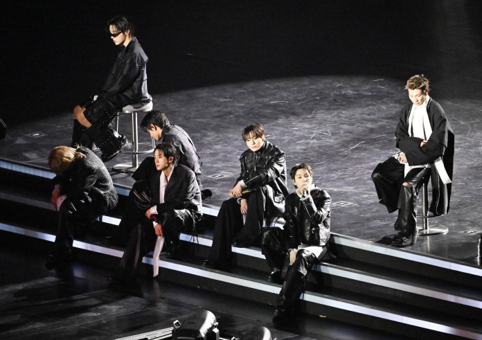 BTS dressed in black outfits seated on steps during a concert, with a spotlight illuminating them from above.