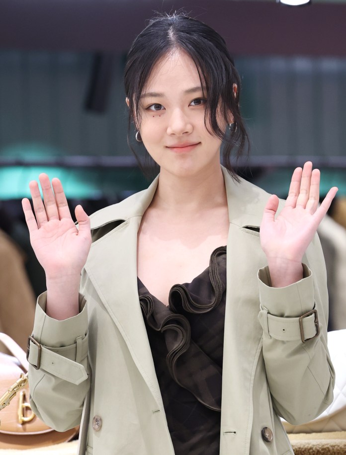 BIBI with black hair wearing a light green trench coat and a black top, smiling and waving at the camera.