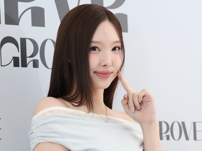 Nayeon with long hair wearing a light blue off-the-shoulder top, smiling and gesturing with her finger near her chin against a light background with stylized text.
