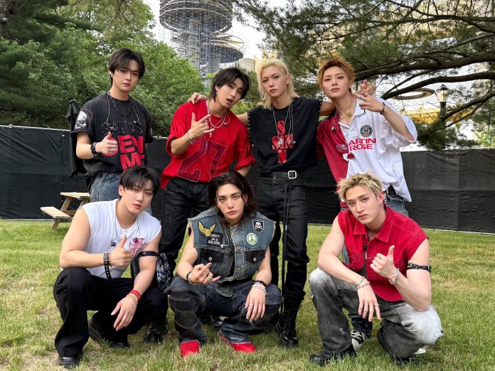 Stray Kids posing for a photo outdoors, dressed in stylish and casual attire, with a mix of red, black, and white clothing. They are arranged in two rows on a grassy area, with trees in the background.