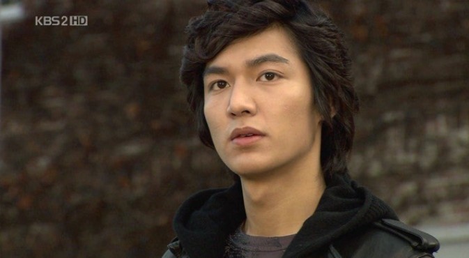 Lee min-ho man with dark, tousled hair and a serious expression, wearing a black hoodie and leather jacket, against a blurred background.