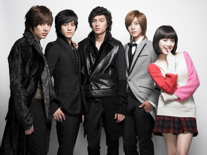 A group of five young adults posing together in stylish outfits, with two males in formal wear, one in a casual black ensemble, and a female in a colorful sweater.