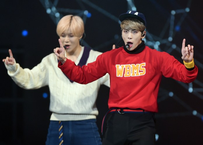 Taemin and Jonghyun on stage, one with pink hair wearing a white sweater and the other with blonde hair in a red sweatshirt, both engaged in a dance pose.