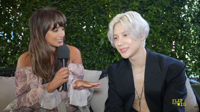 A woman with long brown hair interviews Teamin with silver-white hair, both seated on a couch in front of green foliage. The woman holds a microphone and gestures with her hand while the man smiles.