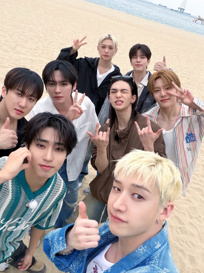 Stray Kids posing together on a beach, smiling and making playful gestures. The sand and ocean can be seen in the background.