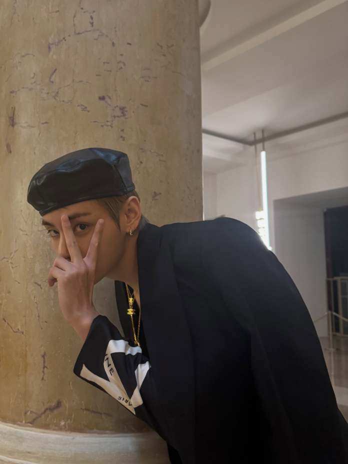 V wearing a leather beret and a stylish black outfit leans against a textured column, making a peace sign with their fingers.