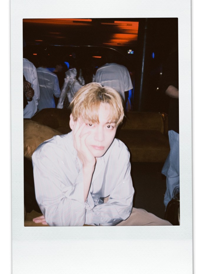 V with blonde hair is sitting at a party, resting their chin on their hand, wearing a light-colored shirt. The background features soft, colorful lighting and blurred figures in a social setting.