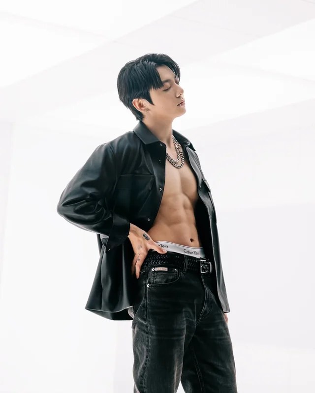 Jungkook poses confidently in a black leather shirt and dark jeans, showcasing his toned physique and accessorized with a chain necklace.
