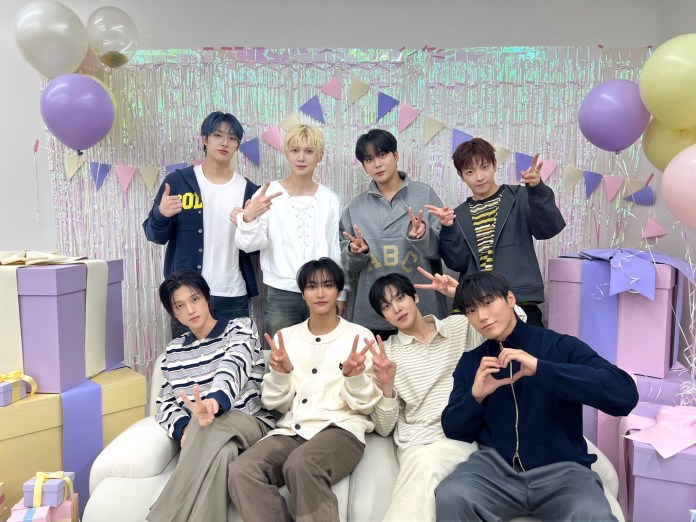 ATEEZ posing together in a festive setting, surrounded by balloons and decorative banners. They are seated and standing in two rows, smiling and making peace signs. Colorful gift boxes are placed nearby.