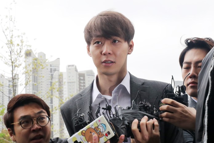 A young man in a gray suit is surrounded by a group of individuals, appearing concerned or anxious, as they navigate a crowded urban setting. Some members of the group are holding recording devices.