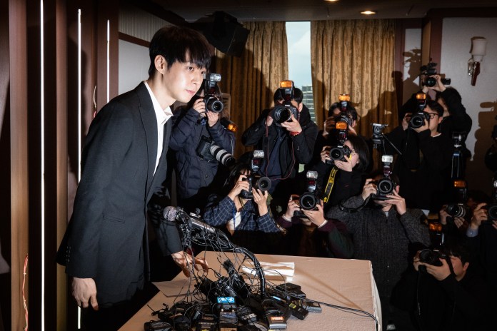 A man in a suit stands at a table surrounded by a crowd of photographers with cameras, capturing the moment during a press event.