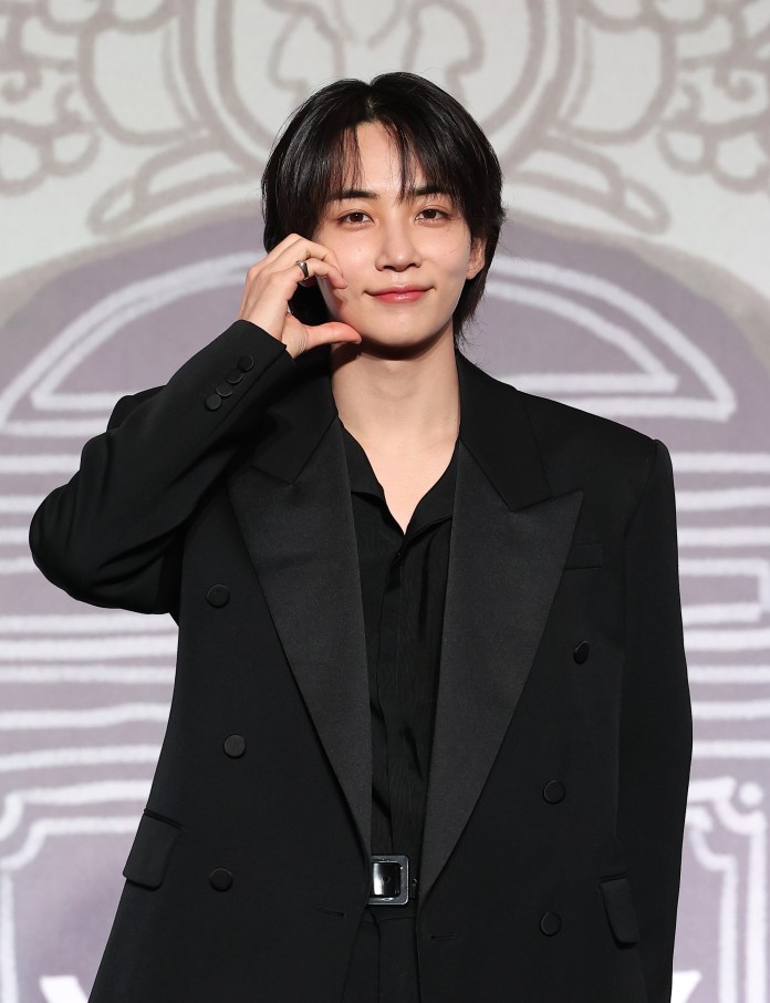 Jeonghan in a black suit and shirt is smiling and making a heart gesture with their hand.