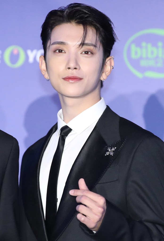 Joshua in a black tuxedo with a white shirt and black tie, smiling and making a hand gesture, at an event backdrop.