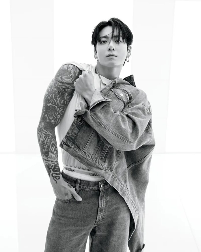 Jungkook displaying a stylish pose while wearing a denim jacket and jeans, showcasing intricate body tattoos. The image is in black and white, emphasizing the fashion statement.