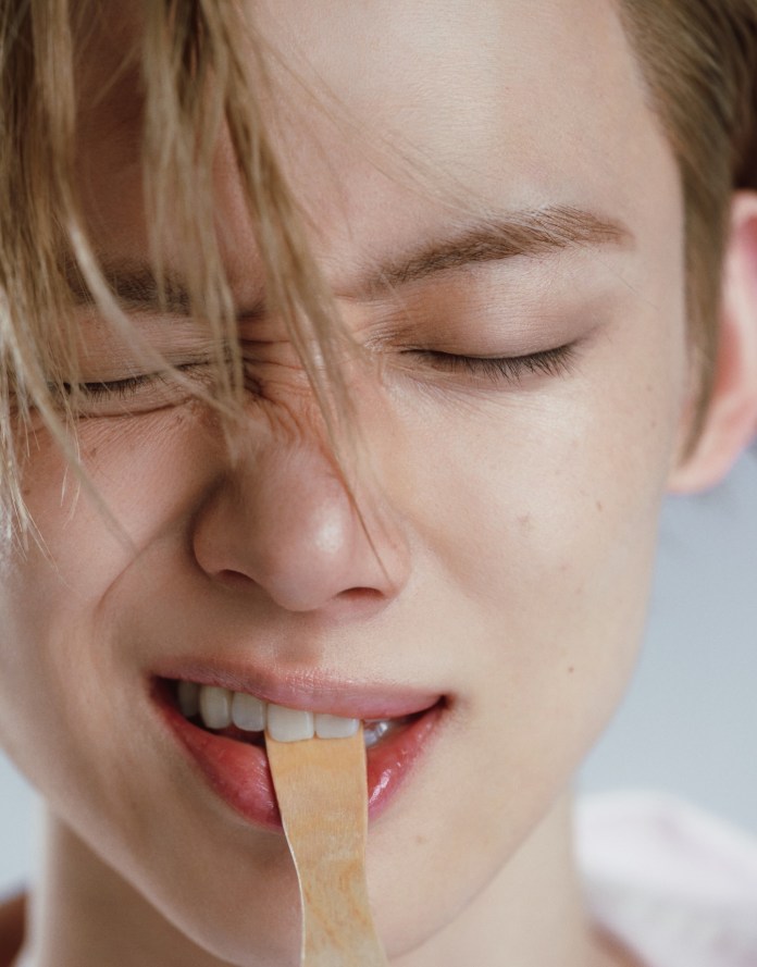 Close-up of Yeonjun with light hair, eyes closed, biting down on a wooden spatula, displaying a focused expression.