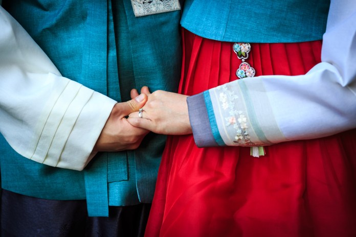 Close-up of two people holding hands, wearing traditional Korean hanbok attire with a turquoise top and red skirt.