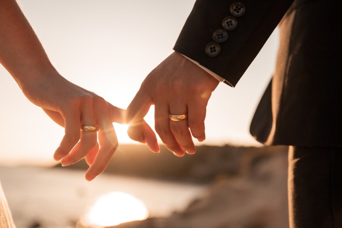 Couple holding hands with wedding rings, backlit by a sunset over the water.