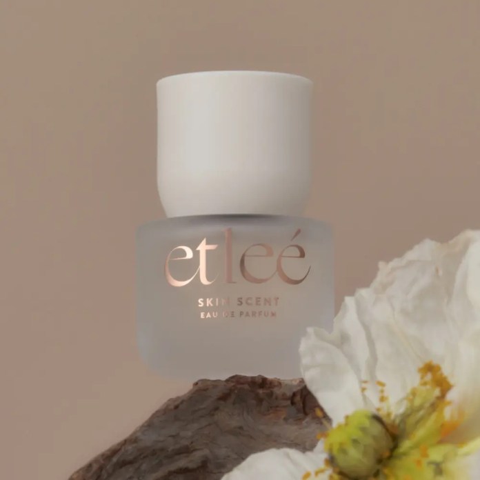 A frosted glass perfume bottle with a white cap, labeled 'etlee SKIN SCENT Eau de Parfum', placed on a piece of wood next to a white flower.