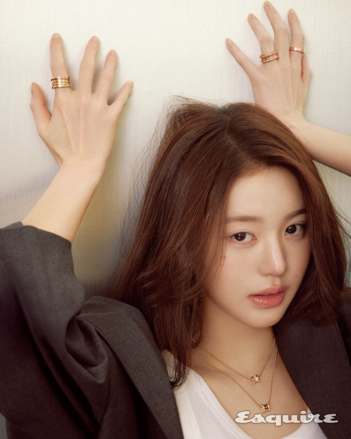 A close-up portrait of Jang Wonyoung with long brown hair, wearing layered necklaces and stylish rings, posed against a light background with her hands positioned on either side of her head.