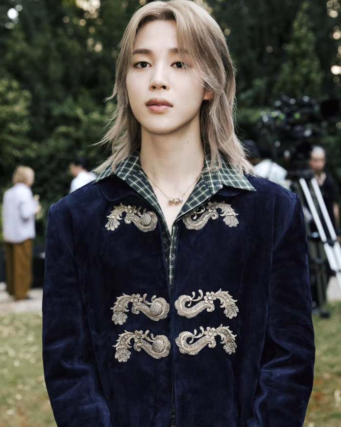 Jimin with long, light hair wearing a blue embroidered jacket over a green checkered shirt, standing outdoors with greenery in the background.