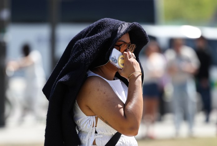 A person wearing a white outfit and dark sunglasses holds a phone to their ear while shielding their face with a dark cloth, in a crowded outdoor setting.