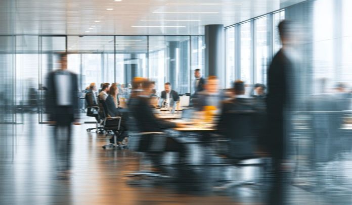 A blurred image of a modern office environment with people engaged in discussions around a conference table.