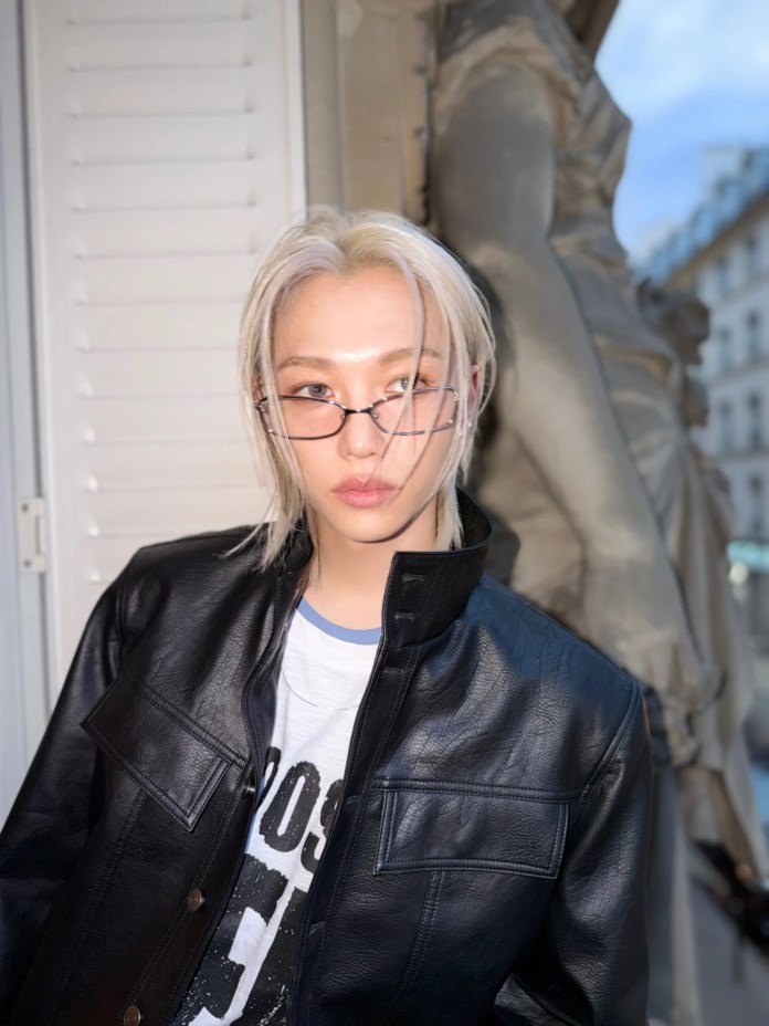 Felix with long platinum blonde hair wearing glasses and a black leather jacket, posed in front of a window with a statue in the background.