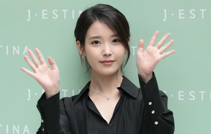 IU in a black shirt waving with both hands, smiling at the camera. The background features the brand name 'J. ESTINA' in light lettering.