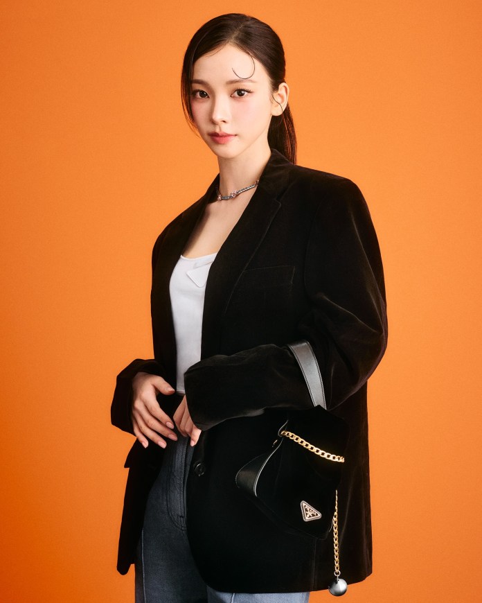 Karina with sleek black hair wearing a black velvet blazer and holding a small handbag, against an orange background.