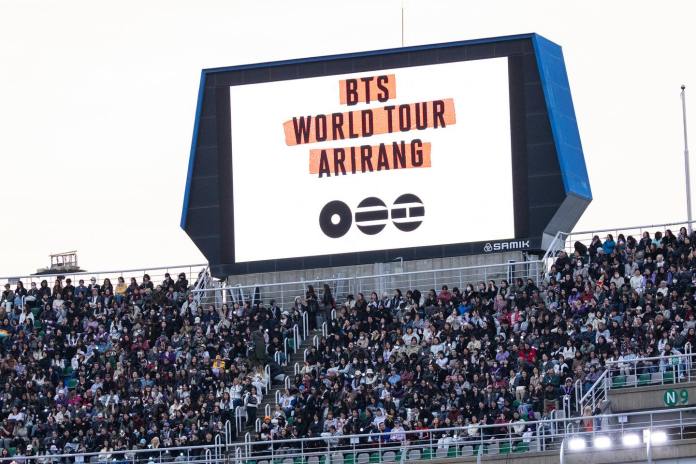 A large digital screen displays the text 'BTS WORLD TOUR ARIRANG' above a crowd of enthusiastic fans in an outdoor stadium setting.